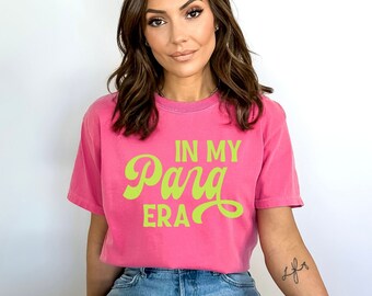 May include: A woman wearing a pink t-shirt with the text "In My Para Era" in green lettering. She is wearing blue denim jeans with rips in the knees.