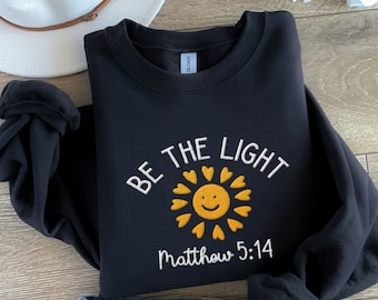 Op de afbeelding: Zwart sweatshirt met witte tekst die "Be the Light" luidt met een gele zonafbeelding en de tekst "Matthew 5:14".