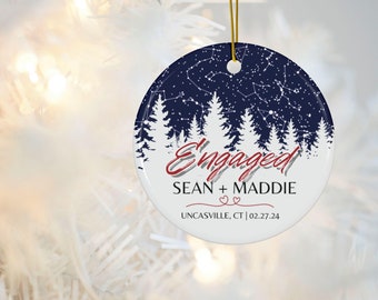 May include: A white ceramic Christmas ornament with a blue background featuring a starry night sky with white pine trees and the text "Engaged Sean + Maddie Uncasville, CT | 02.27.24" in red.