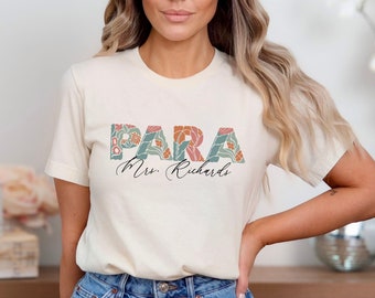 May include: Cream-colored t-shirt with the word "PARA" in floral letters, and the name "Mrs. Richards" in script. The shirt is paired with denim shorts. The text "Natural Sand" is at the bottom.
