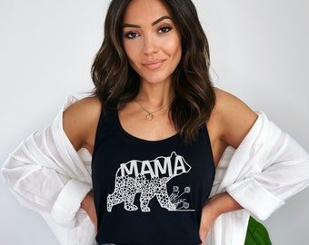 May include: A black tank top with a white floral bear design and the word "MAMA" inside the bear. The model is wearing the tank top with denim shorts and a white cardigan.