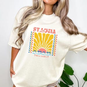May include: Ivory-colored t-shirt with a retro graphic design featuring the text "ST. LUCIA" in red, a yellow sunburst, and blue and pink waves. The design is framed by a checkered border. The text "West Indies" is below.