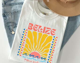 Könnte beinhalten: Ein weißes T-Shirt mit dem Wort "BELIZE" in korallfarbenen Buchstaben über einem gelben Sonnendesign. Die Sonne ist von einem karierten Muster in Blau, Koralle und Gelb umrahmt. Unter der Sonne befinden sich blaue Wellen und der Text "West Indies."