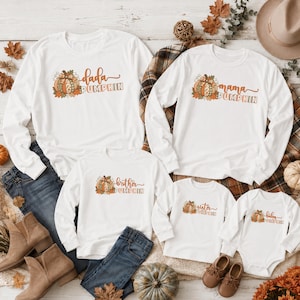 May include: A set of five white long-sleeve shirts and one white long-sleeve bodysuit featuring autumn-themed pumpkin graphics and text including Dada Pumpkin, Mama Pumpkin, Brother Pumpkin, Sister Pumpkin, and Baby Pumpkin in orange and brown.