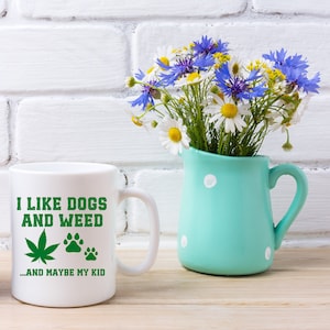 May include: White ceramic mug with the text "I LIKE DOGS AND WEED ...AND MAYBE MY KID" in green. The mug features a green cannabis leaf and paw prints. A turquoise pitcher with white polka dots holds a bouquet of daisies and blue cornflowers.