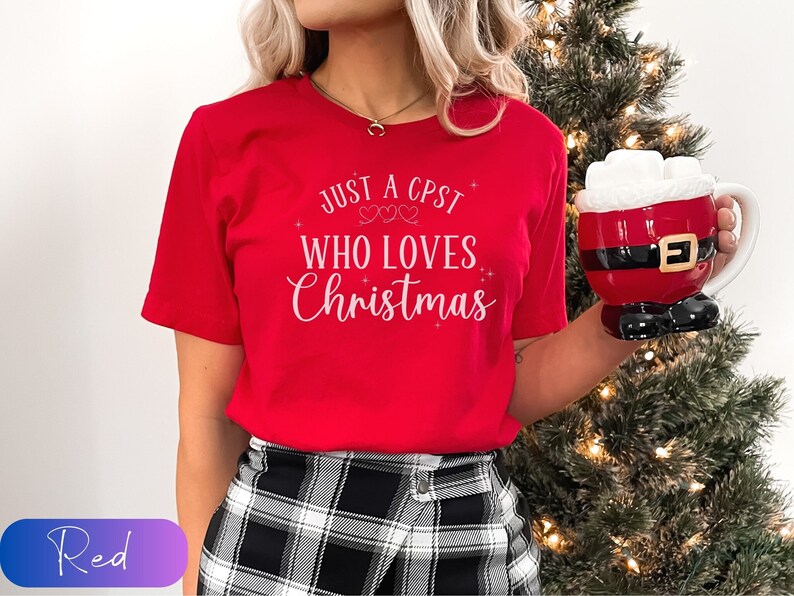 Puede incluir: Camiseta roja con el texto "Just a CPST who loves Christmas" en letras blancas. La camiseta la lleva puesta una persona que sostiene una taza roja de Pap&aacute; Noel.