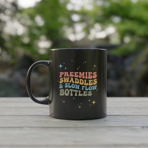 Op de afbeelding: Zwarte koffiemok met een wit ontwerp met de tekst "Preemies Swaddles & Slow Flow Bottles" met regenboogkleurige letters en sterren.