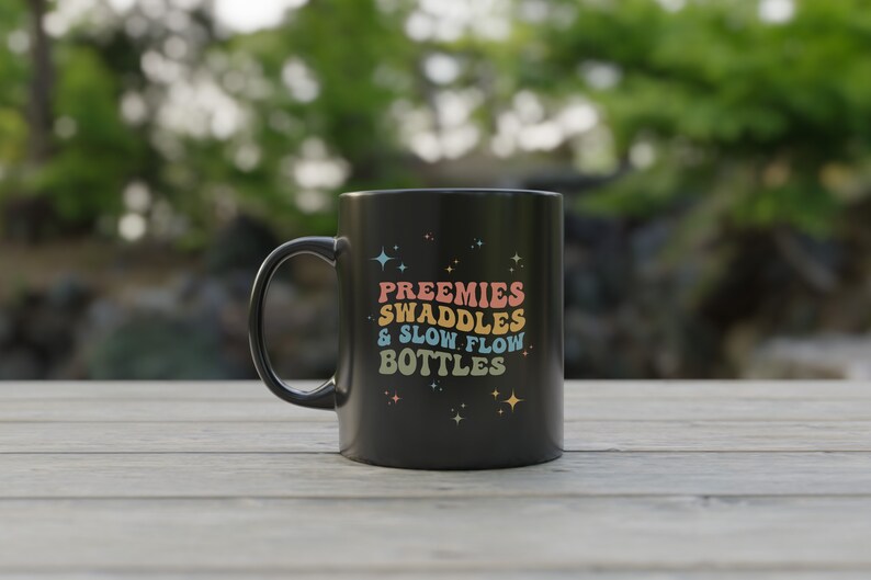 Op de afbeelding: Zwarte koffiemok met een wit ontwerp met de tekst "Preemies Swaddles & Slow Flow Bottles" met regenboogkleurige letters en sterren.