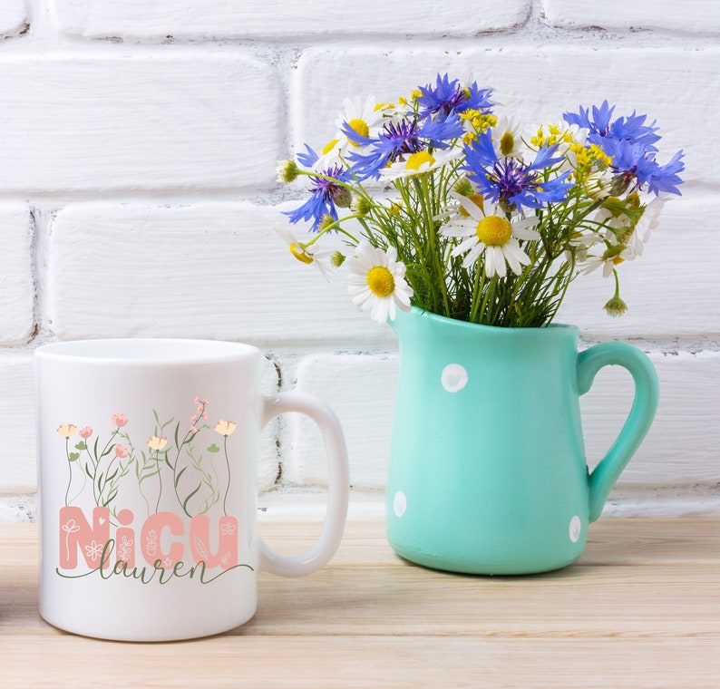 May include: White ceramic mug with a pink floral design and the text "NICU Lauren". The mug is sitting on a wooden surface next to a light blue pitcher with white polka dots filled with a bouquet of blue and white flowers.