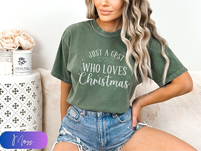 Puede incluir: Una camiseta verde con el texto "Just a CPST who loves Christmas" en letras blancas. La camiseta tiene un aspecto desgastado y la lleva puesta una persona.