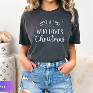 Puede incluir: Una camiseta gris oscuro con el texto "Just a CPST who loves Christmas" en letras blancas. La camiseta tiene cuello redondo y mangas cortas.