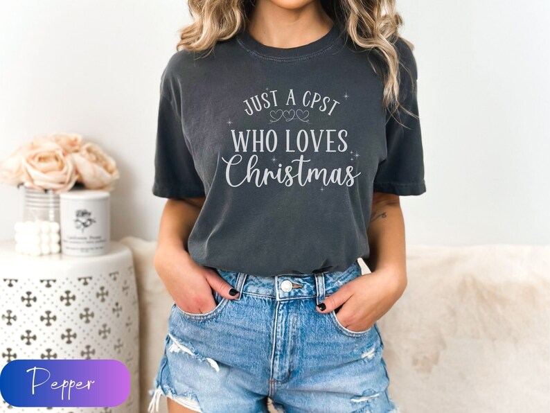 Puede incluir: Una camiseta gris oscuro con el texto "Just a CPST who loves Christmas" en letras blancas. La camiseta tiene cuello redondo y mangas cortas.