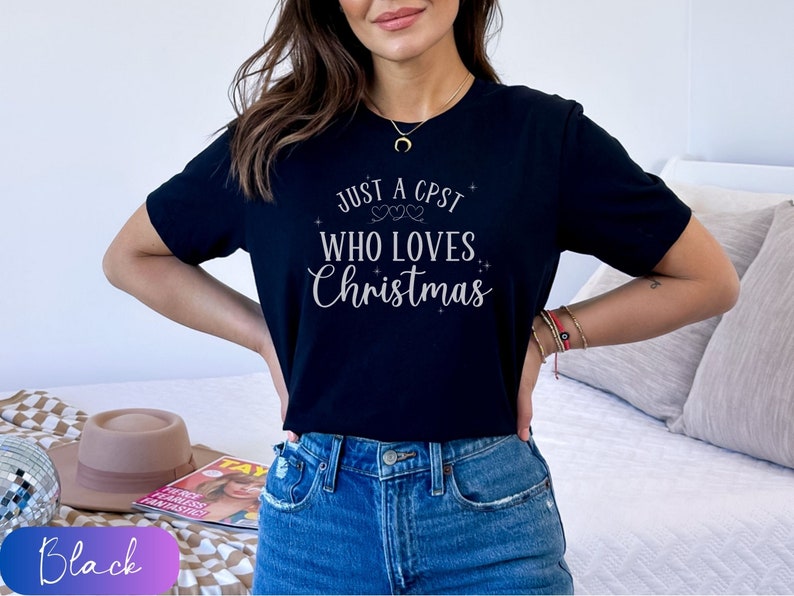 Puede incluir: Una camiseta negra con el texto "Just a CPST who loves Christmas" en letras plateadas brillantes.