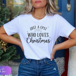 Puede incluir: Camiseta blanca con el texto "Just a CPST who loves Christmas" en letras negras con dos peque&ntilde;os corazones sobre la palabra "loves".