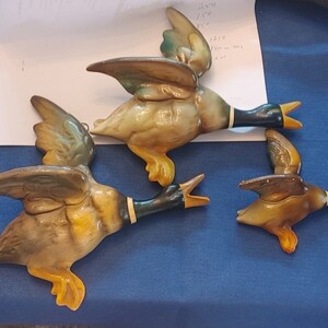 Peut inclure: Trois figurines de canard en céramique en vol, peintes dans des tons de brun, jaune et vert. Les canards ont des yeux et des becs noirs.