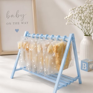 May include: A light blue wooden A-frame display stand featuring a horizontal bar with hanging rings. Several translucent white plastic bags with a white polka dot pattern are suspended from the rings, each containing a light-colored baked food item.
