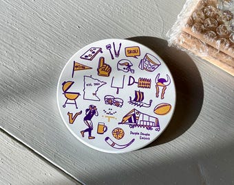May include: A white round coaster with a purple and yellow design featuring various Minnesota Vikings themed illustrations, including a football helmet, a Viking ship, a grill, a beer mug, and the text "Purple People Eaters".