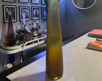 Könnte beinhalten: Eine hohe, schlanke Glasvase in Bernsteinfarbe mit verjüngtem Hals und breitem, kreisförmigem Boden. Das Glas hat eine glatte, durchscheinende Oberfläche, die von einem tiefen, gesättigten Braun am Boden zu einem helleren Goldton oben übergeht.