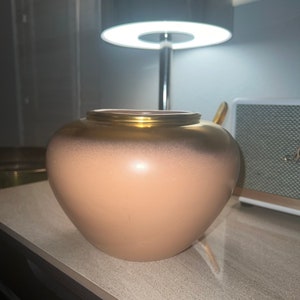 May include: A light pink ceramic vase with a gold rim. The vase has a round, slightly oval shape.