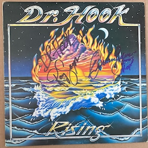 Puede incluir: Portada del álbum "Rising" de Dr. Hook. La obra de arte presenta un amanecer estilizado sobre las olas del océano, con llamas y una luna creciente. El título del álbum y el nombre de la banda se muestran en una fuente metálica, y la portada está firmada.