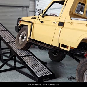 Puede incluir: Un camión de juguete amarillo sube por una rampa de metal negro. La rampa tiene una superficie perforada. El camión tiene neumáticos grandes y un diseño detallado. La imagen está ambientada en un garaje.