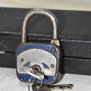 May include: A vintage blue and silver padlock with a key inserted and another key beside it. The lock has a curved metal handle and the words "MADE IN BULGARIA" are visible. The lock is on a white surface.