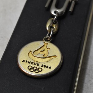Könnte beinhalten: Ein goldfarbener Schlüsselanhänger mit einem runden Anhänger mit dem Logo der Olympischen Spiele von Athen 2004. Der Anhänger hat ein stilisiertes Bild und den Text "ATHENS 2004" in Schwarz. Der Schlüsselanhänger hat einen silberfarbenen Verschluss und eine Kette.