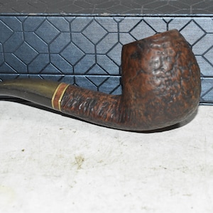 May include: A brown tobacco pipe with a curved stem and a textured bowl. The stem has a brass band with a thin red stripe. The pipe is set against a blue patterned background.