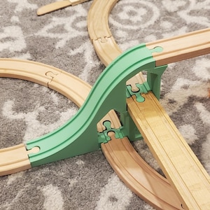 Puede incluir: Un set de vías de tren de juguete de madera con un puente verde, un puente rojo y naranja y un vagón azul. La vía está colocada sobre una alfombra gris.
