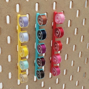 May include: Three wall-mounted thread organizers with colorful spools of thread. Each organizer has six spools of thread in different colors, including pink, purple, blue, yellow, and red.