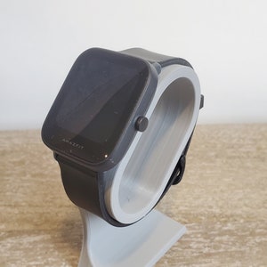 May include: A black smartwatch with a gray stand. The watch face displays the word "AMAZFIT".
