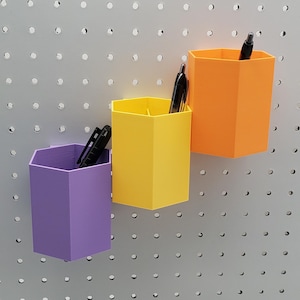May include: Three hexagonal pencil holders in purple, yellow, and orange, mounted on a pegboard wall. Each holder has a black pen inside.