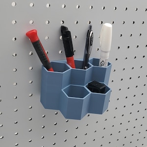 Pode incluir: Um porta-canetas hexagonal azul com vários utensílios de escrita. O suporte é montado em um painel perfurado cinza. Os itens incluem uma chave de fenda vermelha, canetas pretas e um marcador branco.