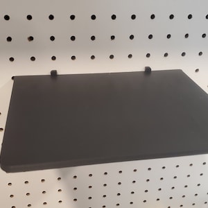 Puede incluir: Un estante negro con dos soportes metálicos fijados a una pared de tablero perforado blanco.