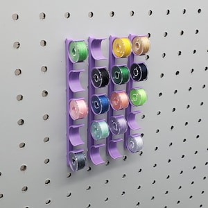 May include: A purple plastic thread organizer with 20 slots for holding spools of thread. The organizer is mounted on a pegboard wall. The spools of thread are in various colors, including green, blue, yellow, and pink.