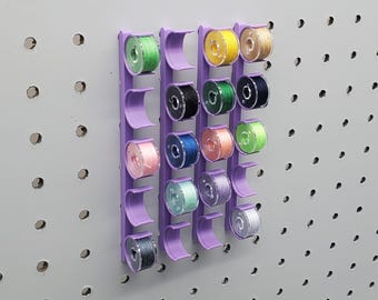 Puede incluir: Un organizador de hilo de plástico morado con 20 ranuras para guardar carretes de hilo. El organizador está montado en una pared de tablero perforado. Los carretes de hilo son de varios colores, incluyendo verde, azul, amarillo y rosa.