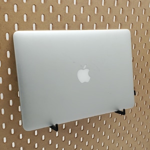 以下が含まれることがあります： 黒いアップルのロゴが付いた銀色のラップトップが、黒い金属製のフックを使ってペグボードの壁に取り付けられています