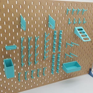 Puede incluir: Un tablero de clavijas con varios ganchos, estantes y organizadores de pl&aacute;stico azul turquesa. El tablero de clavijas est&aacute; hecho de madera y tiene un acabado marr&oacute;n.