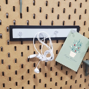 Op de afbeelding: Een wit prikbord met een zwarte metalen strip met drie haken. Een paar witte oordopjes hangen aan de haken. Een groene kaart met een kerstboomontwerp en de tekst "Prettige Kerst" is vastgemaakt aan het prikbord. Een kleine groene potplant staat op een groen plankje dat aan het prikbord is bevestigd.