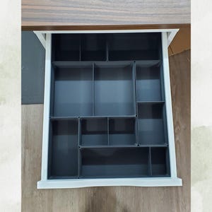 May include: A white-framed drawer with a dark gray interior, divided into multiple compartments of varying sizes. The drawer is open, revealing the organizational system within, designed for storage and tidiness.