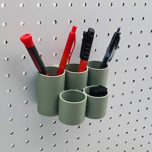 Puede incluir: Un organizador de pared de plástico verde con seis compartimentos que contiene cuatro bolígrafos y un objeto pequeño. Dos de los bolígrafos son rojos y dos son negros.