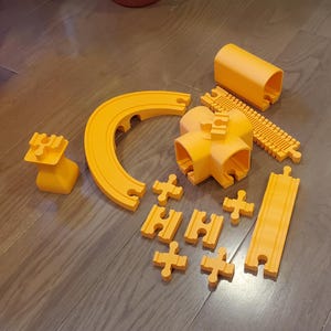 Puede incluir: Un conjunto de piezas de vía de plástico naranja para un tren de juguete. El conjunto incluye vía recta, vía curva y una pieza de túnel. También hay varias piezas de conexión.