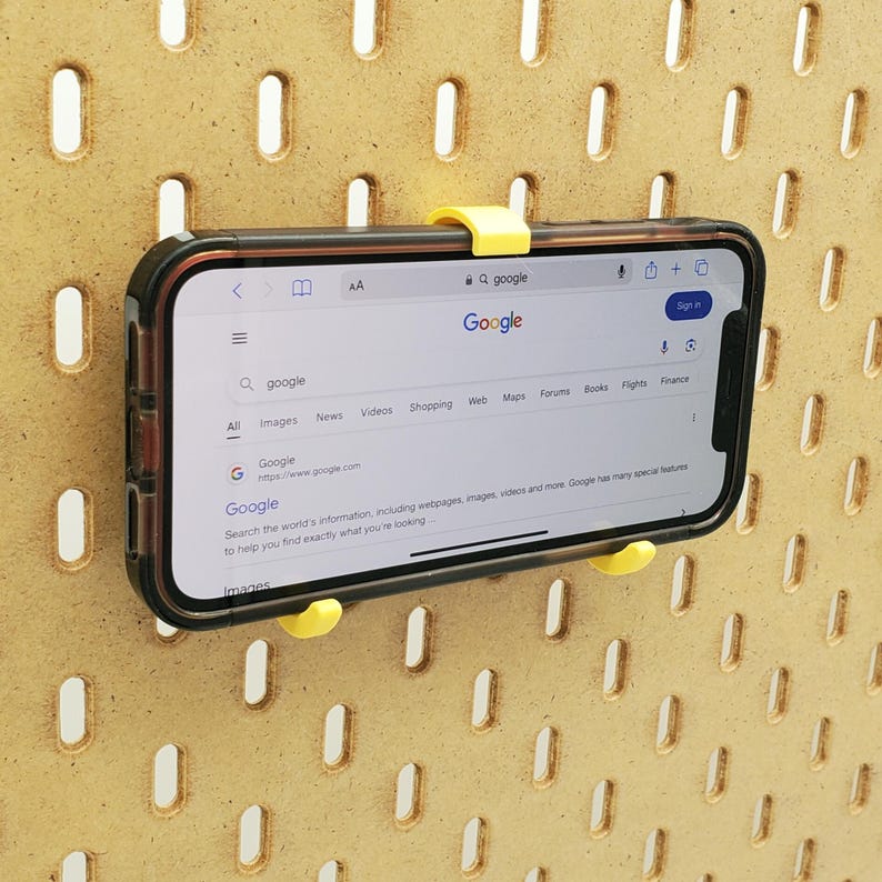 May include: A yellow phone holder with a black smartphone attached to it. The phone is displaying the Google search page with the text "Google" in a large font. The phone holder is attached to a pegboard wall.