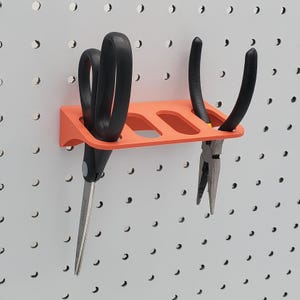May include: Orange plastic tool holder with three slots mounted on a white pegboard wall. A pair of black scissors and two pairs of black pliers are hanging in the slots.