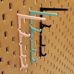 Puede incluir: Tres ganchos de plástico montados en una pared de tablero perforado. Los ganchos son de color rosa, azul claro y negro.