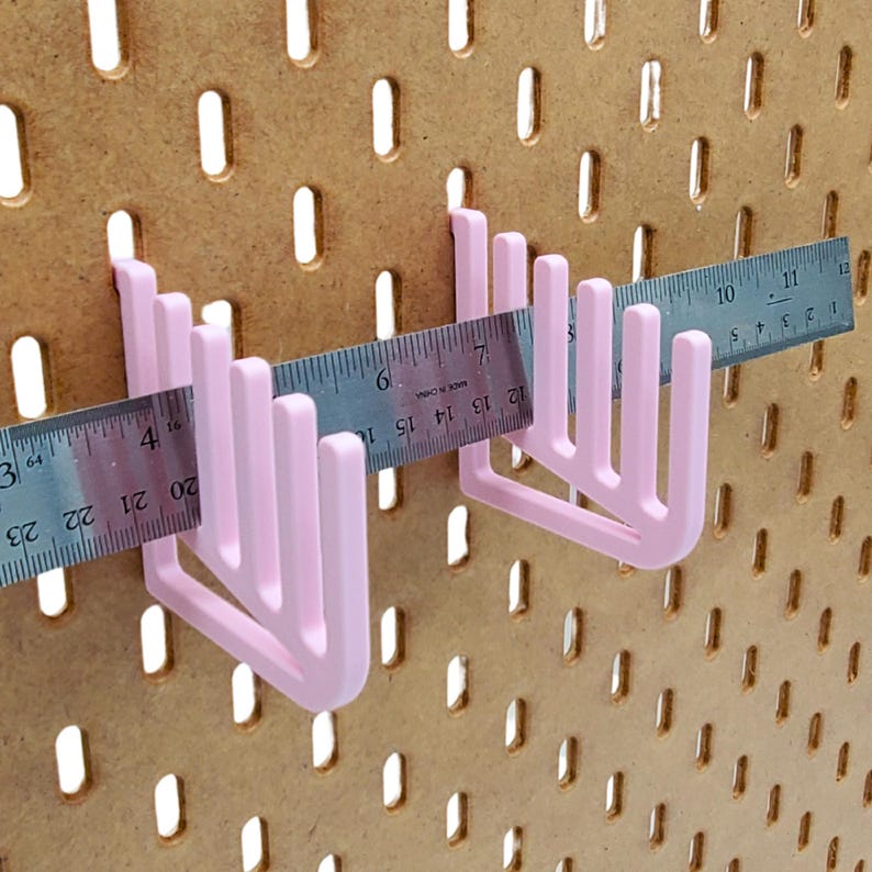 Op de afbeelding: Twee roze plastic pegboard haken, elk met vier tanden, zijn gemonteerd op een bruin pegboard. Een metalen liniaal is over de haken gelegd en meet 17,8 cm.