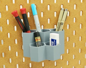 May include: A grey hexagonal organiser with six compartments holds various items, including two red screwdrivers, a blue marker, two black pens, and a white eraser. The organiser is mounted on a pegboard.
