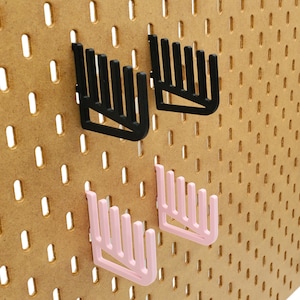 Op de afbeelding: Twee sets zwarte en roze pegboard haken. De haken zijn kamvormig met meerdere tanden. De haken zijn gemonteerd op een bruin pegboard.