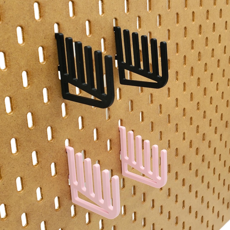 Op de afbeelding: Twee sets zwarte en roze pegboard haken. De haken zijn kamvormig met meerdere tanden. De haken zijn gemonteerd op een bruin pegboard.