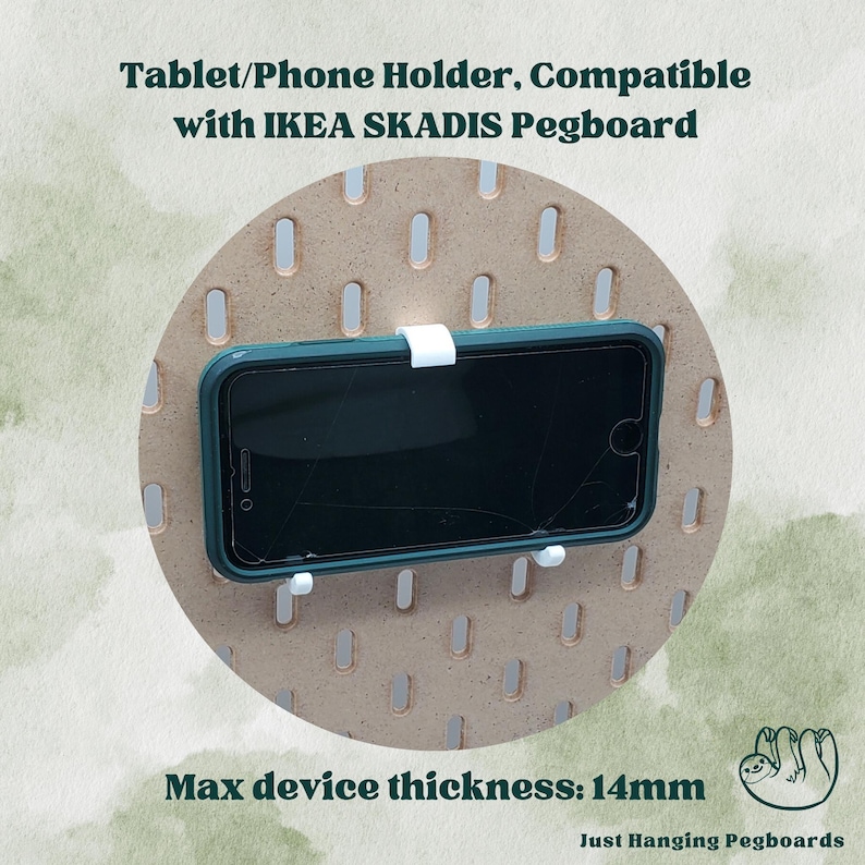 May include: A white phone holder for an IKEA SKADIS pegboard. The holder is made of plastic and has a maximum device thickness of 14mm. The holder is shown holding a black smartphone on a brown pegboard.